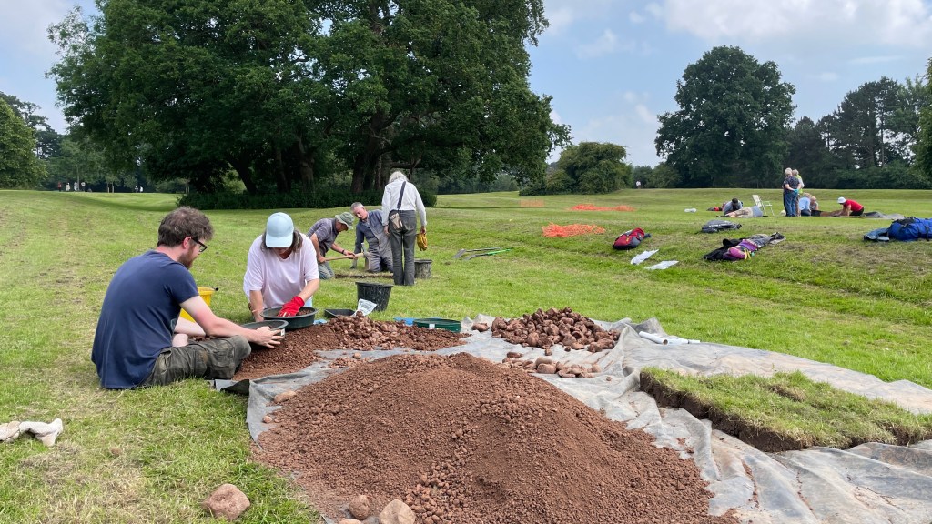 People digging test pits and sieving soil for finds in grass parkland.